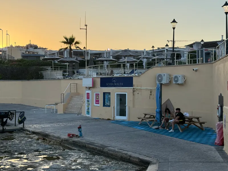 The rocky coast behind Paceville at sunset: a dive shop, picnic tables and the last swimmers of the day. Every stretch of this coastline has a corner like this